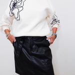 Person wearing a white sweater with black abstract designs and a black leather skirt against a white background
