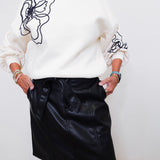 Person wearing a white sweater with black abstract designs and a black leather skirt against a white background