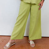 Person wearing a green outfit with wide-leg pants and a woven bag, standing on a terracotta floor.