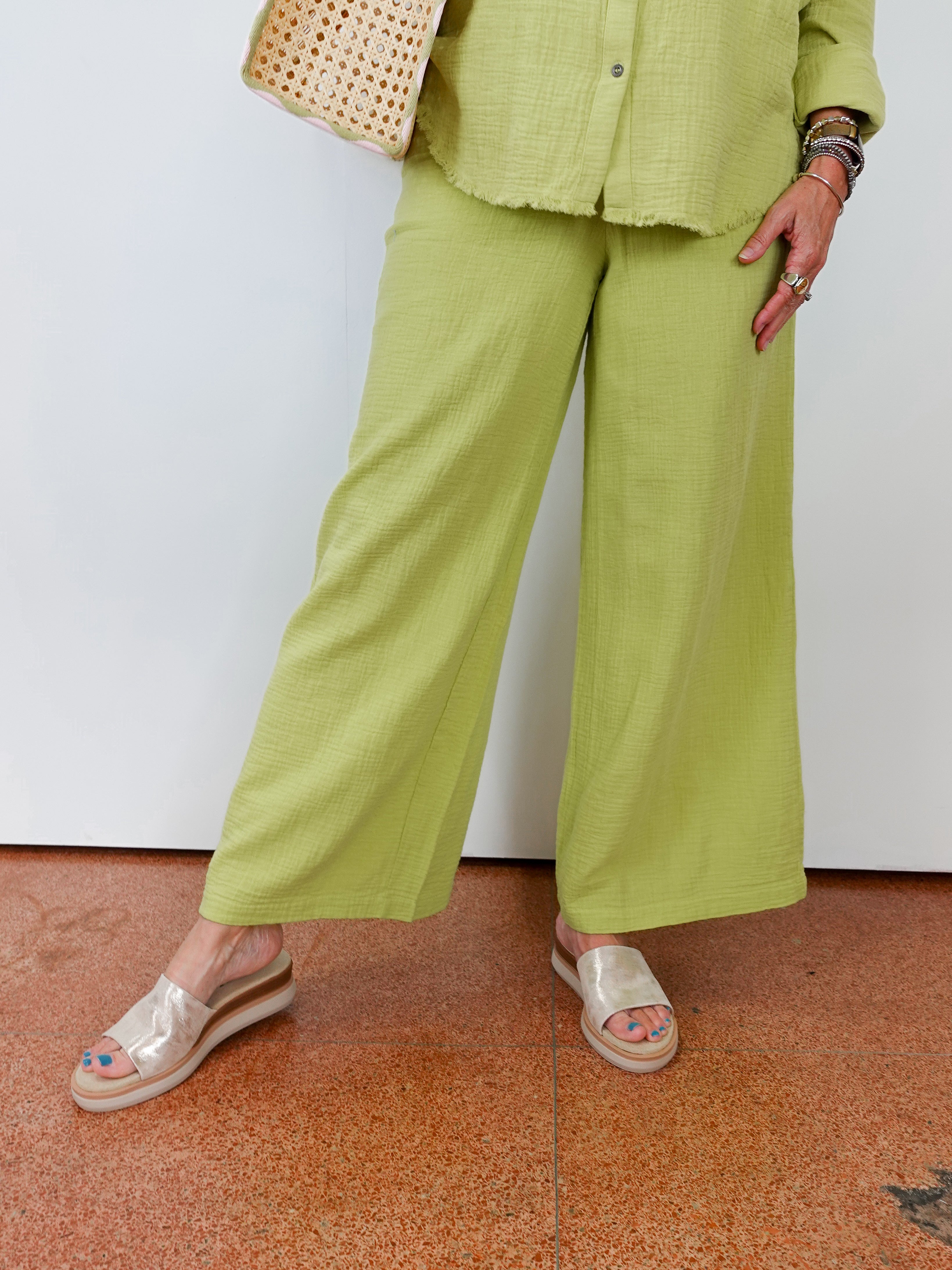 Person wearing a green outfit with wide-leg pants and a woven bag, standing on a terracotta floor.