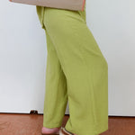 Person wearing green pants and sandals, standing on a brown floor with a white wall in the background.
