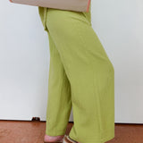 Person wearing green pants and sandals, standing on a brown floor with a white wall in the background.