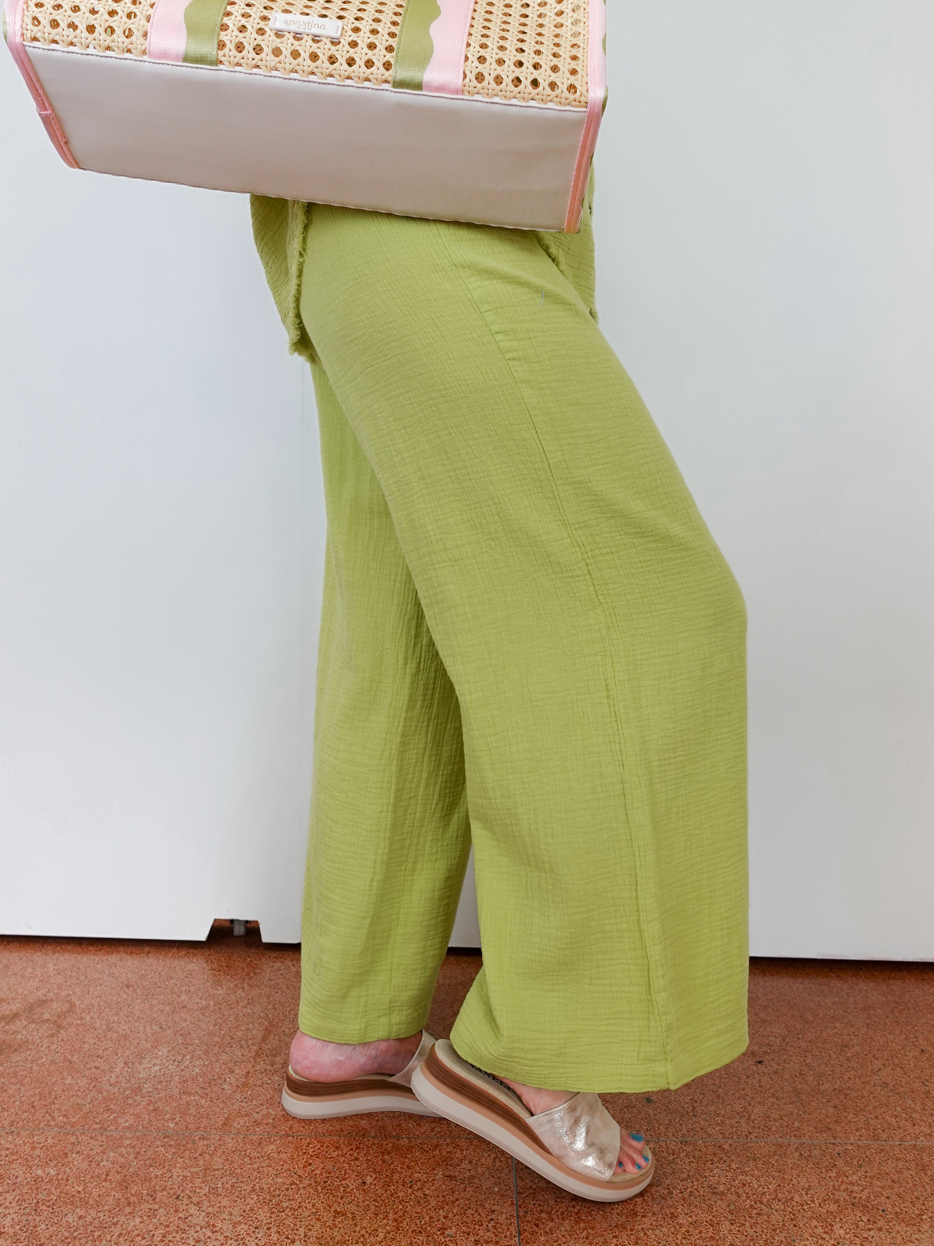 Person wearing green pants and sandals, standing on a brown floor with a white wall in the background.