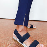 A close-up of a person's feet wearing navy blue strappy espadrille sandals with a white braided sole and a tan wedge heel, paired with navy trousers.