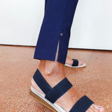 A close-up of a person's feet wearing navy blue strappy espadrille sandals with a white braided sole and a tan wedge heel, paired with navy trousers.