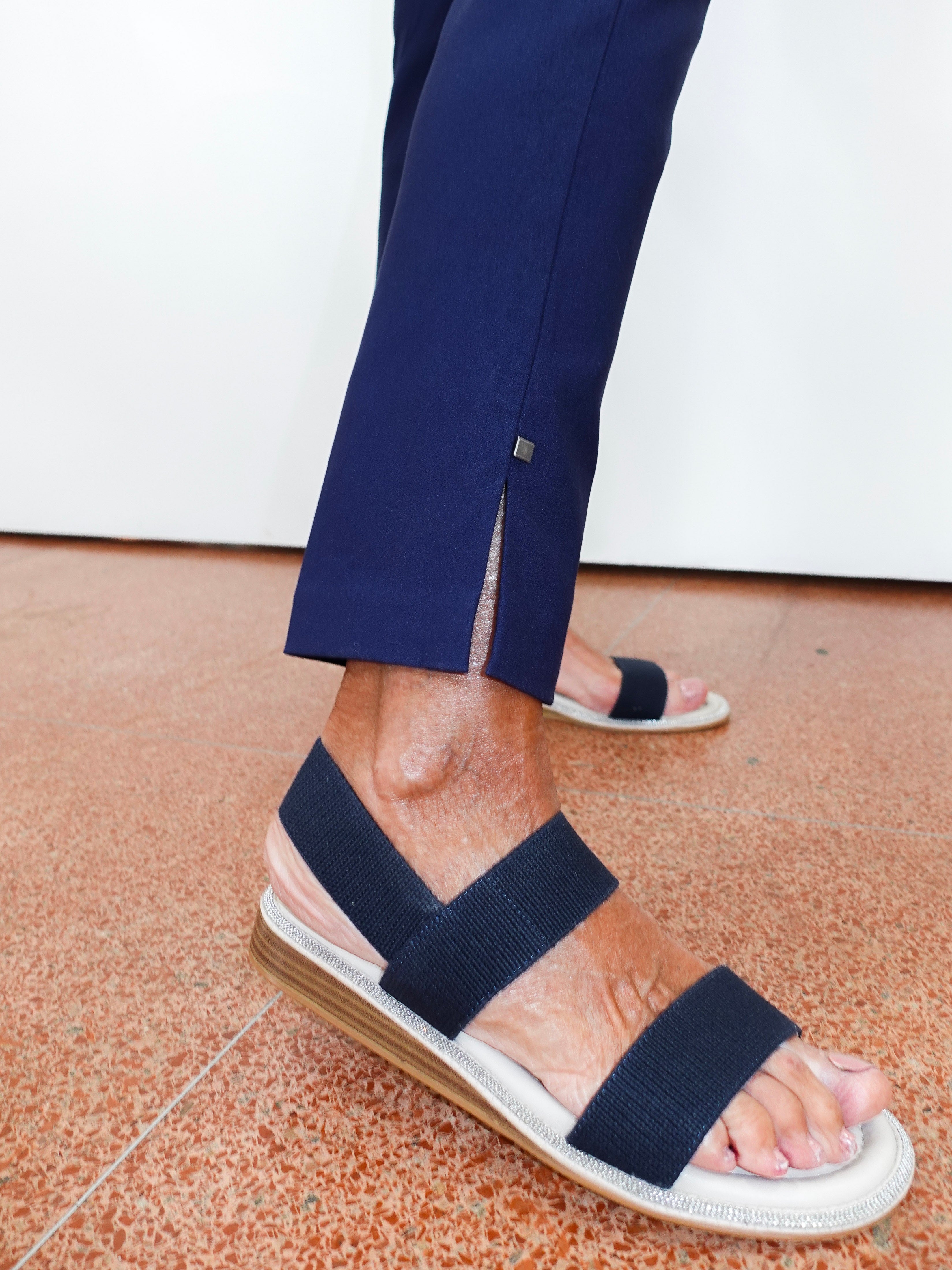 A close-up of a person's feet wearing navy blue strappy espadrille sandals with a white braided sole and a tan wedge heel, paired with navy trousers.