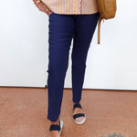 A person wearing navy blue slim-fit trousers, a beige and orange striped blouse, and navy slide sandals, carrying a tan shoulder bag against a white wall.