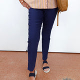 A person wearing navy blue slim-fit trousers, a beige and orange striped blouse, and navy slide sandals, carrying a tan shoulder bag against a white wall.