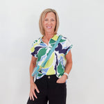 Woman wearing a colorful patterned shirt and black shorts with a green bag against a white background