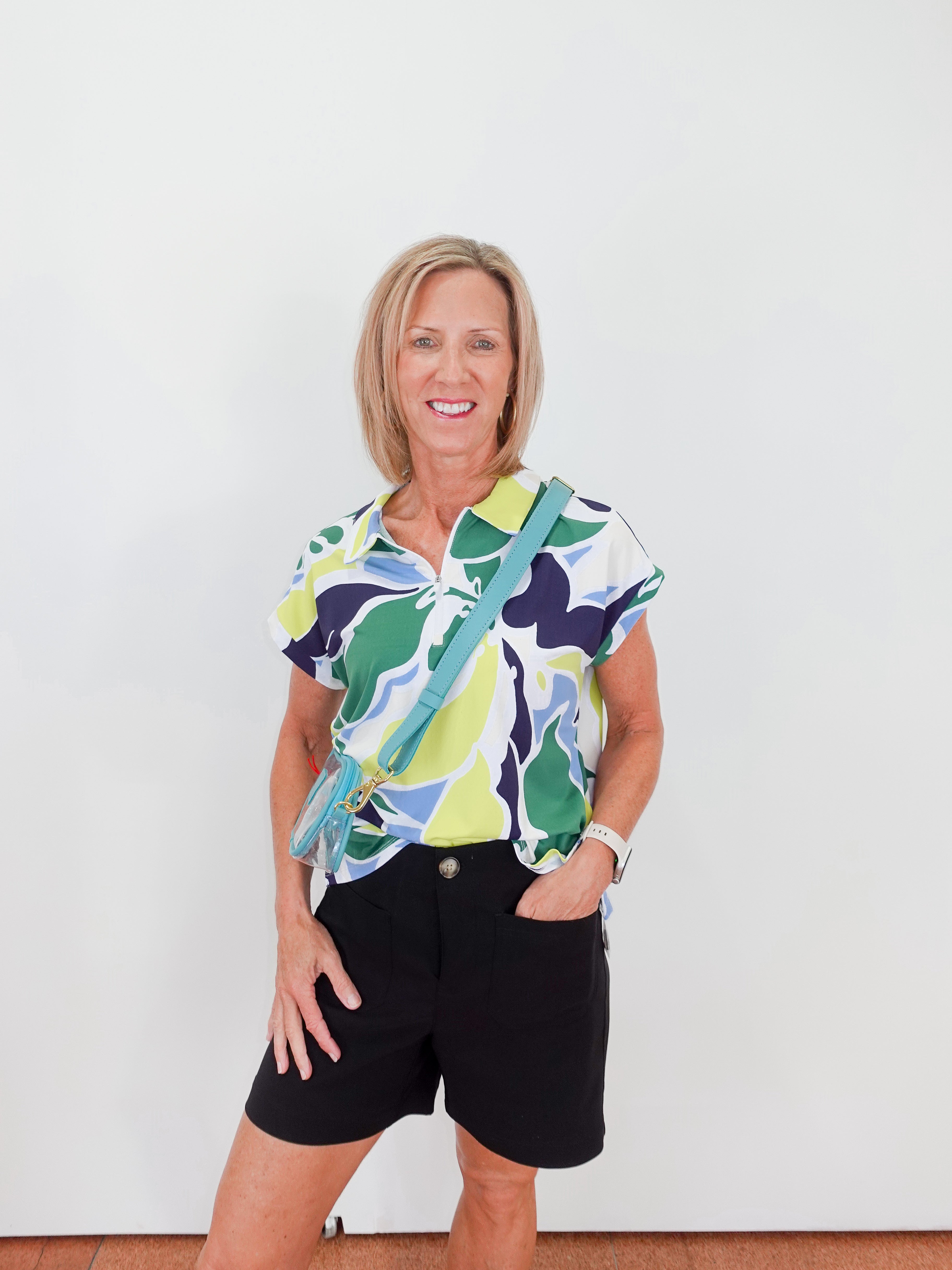 Woman wearing a colorful patterned shirt and black shorts with a green bag against a white background