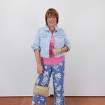 Woman wearing a denim jacket, pink shirt, and blue floral pants against a white wall.