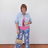 Woman wearing a denim jacket, pink shirt, and blue floral pants against a white wall.