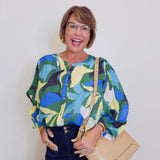 Woman wearing a colorful patterned blouse with a beige handbag against a plain background