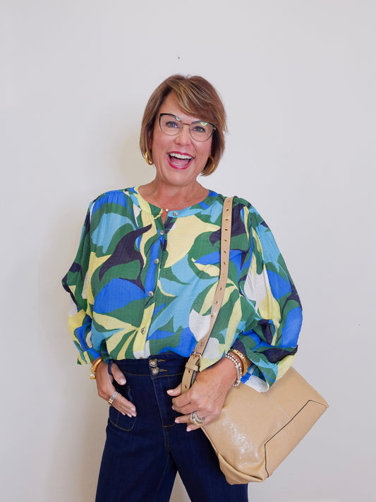 Woman wearing a colorful patterned blouse with a beige handbag against a plain background