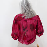A woman with long gray hair is shown from behind wearing a vibrant magenta pink blouse with a dark red abstract floral pattern, paired with light beige pants.