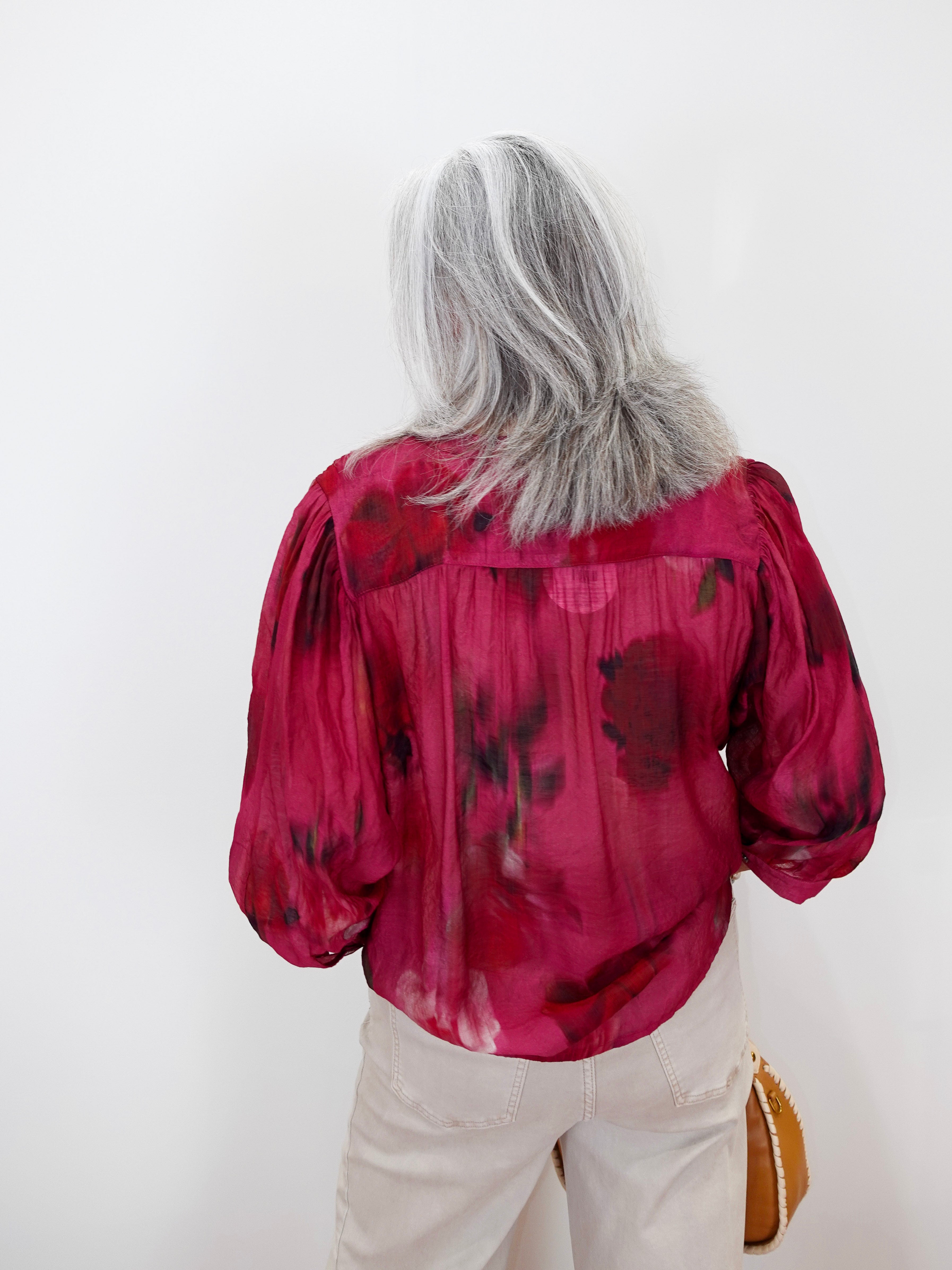 A woman with long gray hair is shown from behind wearing a vibrant magenta pink blouse with a dark red abstract floral pattern, paired with light beige pants.