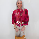 An older woman with gray hair stands against a white wall wearing a vibrant magenta and red abstract print blouse, beige trousers, a white belt with a large gold buckle, and holding a tan leather handbag.
