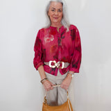 An older woman with gray hair stands against a white wall wearing a vibrant magenta and red abstract print blouse, beige trousers, a white belt with a large gold buckle, and holding a tan leather handbag.