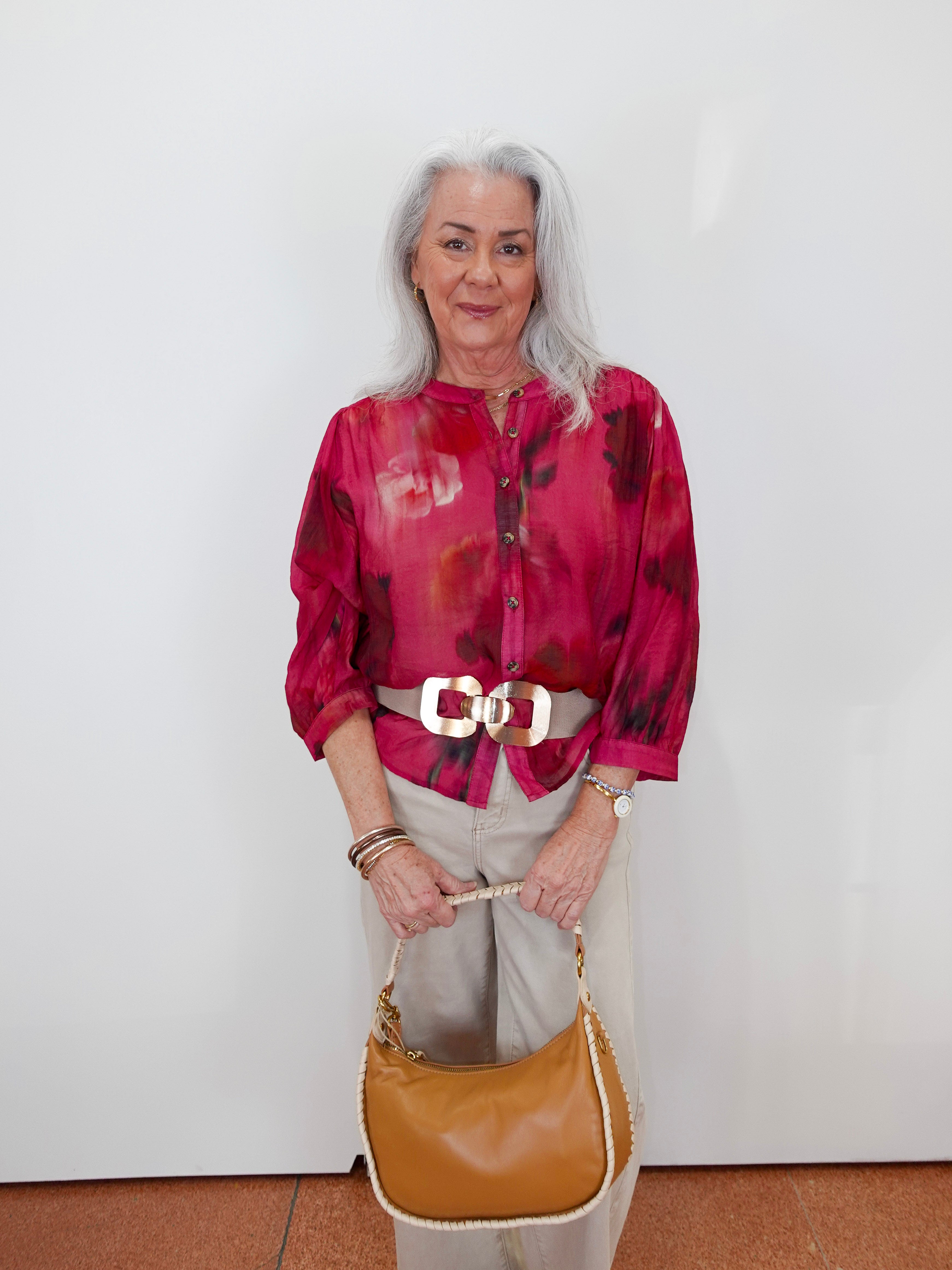 An older woman with gray hair stands against a white wall wearing a vibrant magenta and red abstract print blouse, beige trousers, a white belt with a large gold buckle, and holding a tan leather handbag.