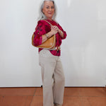 An older woman with gray hair stands against a white wall wearing a bright pink patterned blouse, beige wide-leg pants, and carrying a tan shoulder bag.