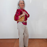 An older woman with gray hair stands against a white wall wearing a bright pink patterned blouse, beige wide-leg pants, and carrying a tan shoulder bag.
