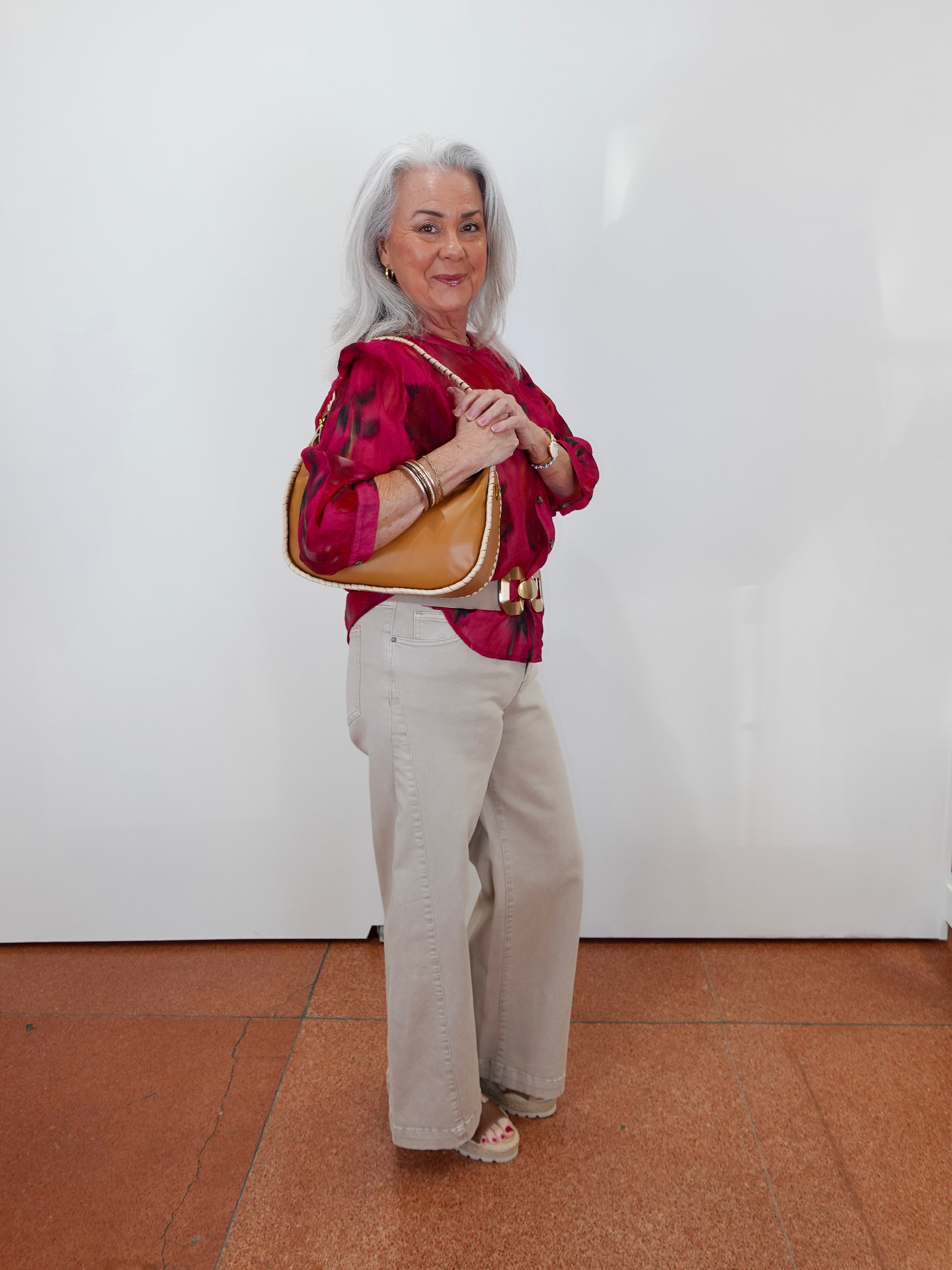 An older woman with gray hair stands against a white wall wearing a bright pink patterned blouse, beige wide-leg pants, and carrying a tan shoulder bag.
