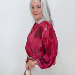 An older woman with long gray hair is shown in profile against a plain white wall, wearing a sheer long-sleeve blouse with a dark red and black abstract print, paired with off-white pants and carrying a tan handbag.