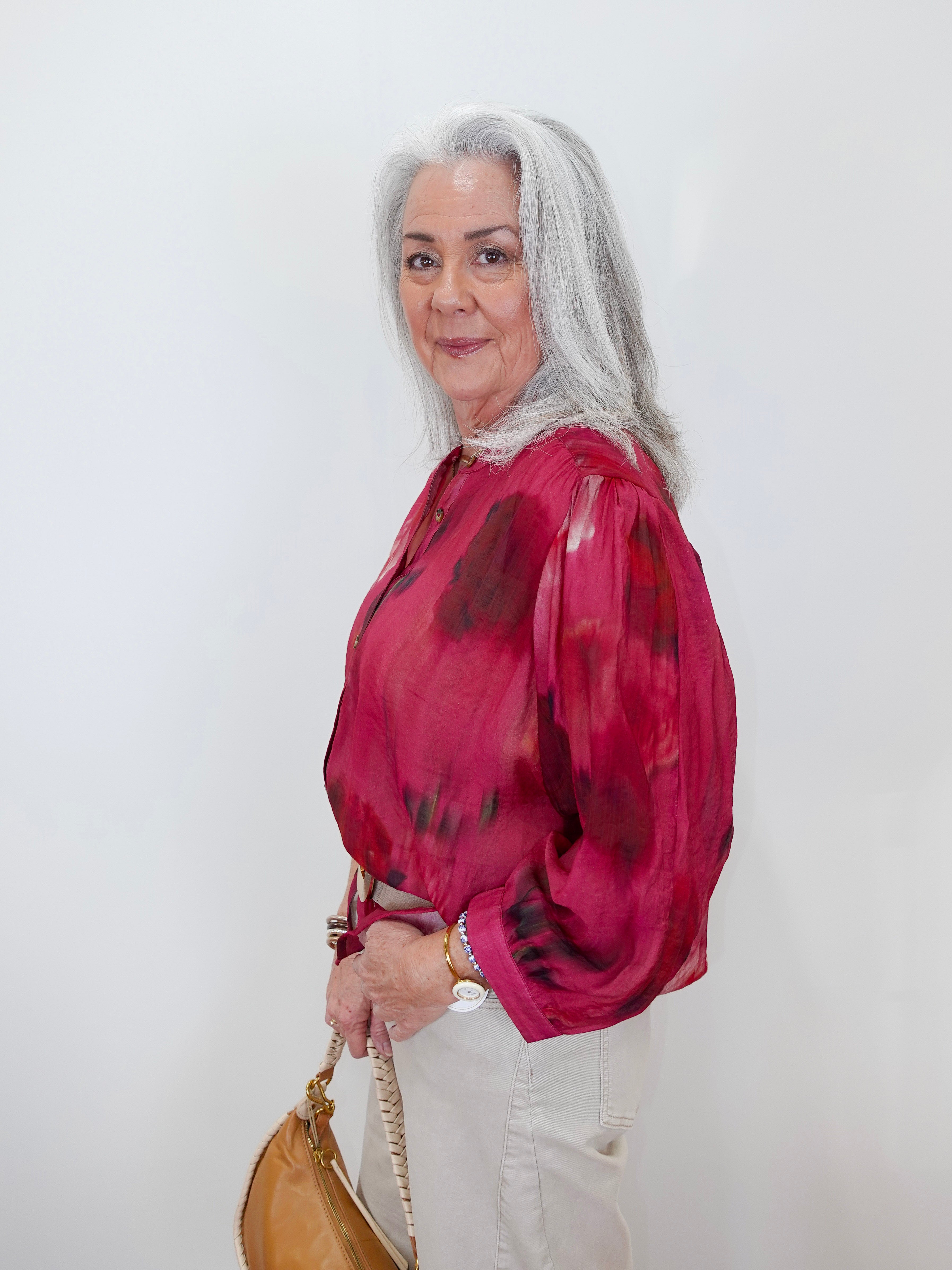 An older woman with long gray hair is shown in profile against a plain white wall, wearing a sheer long-sleeve blouse with a dark red and black abstract print, paired with off-white pants and carrying a tan handbag.
