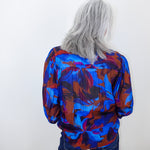 A person with long gray hair seen from behind, wearing a vibrant blue, red, and purple abstract patterned blouse and dark blue jeans, standing against a plain white wall.