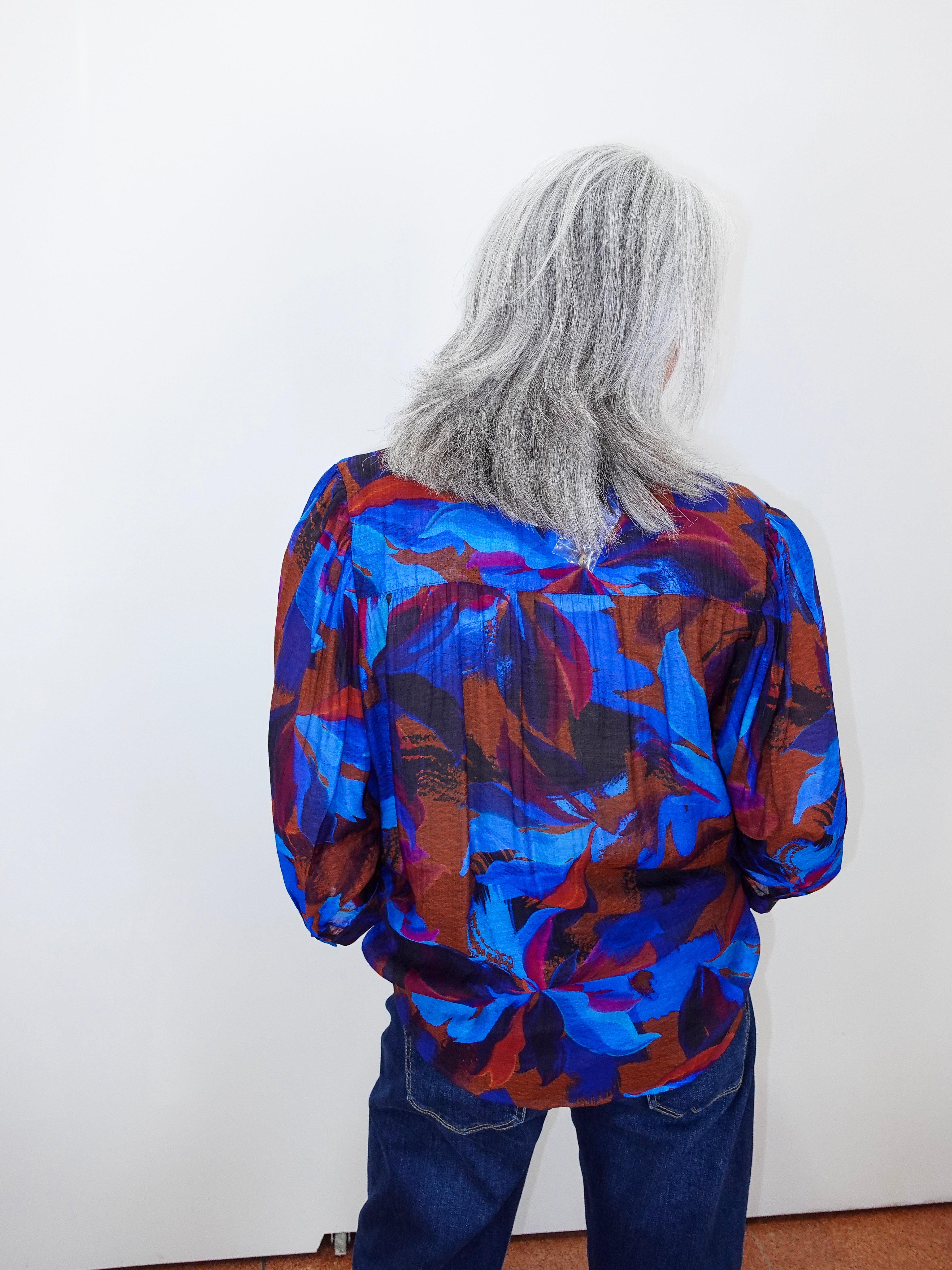 A person with long gray hair seen from behind, wearing a vibrant blue, red, and purple abstract patterned blouse and dark blue jeans, standing against a plain white wall.