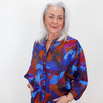 An older woman with silver hair stands against a plain white wall wearing a vibrant, long-sleeved button-up blouse with an abstract floral pattern in blue, purple, and red, paired with dark blue jeans.