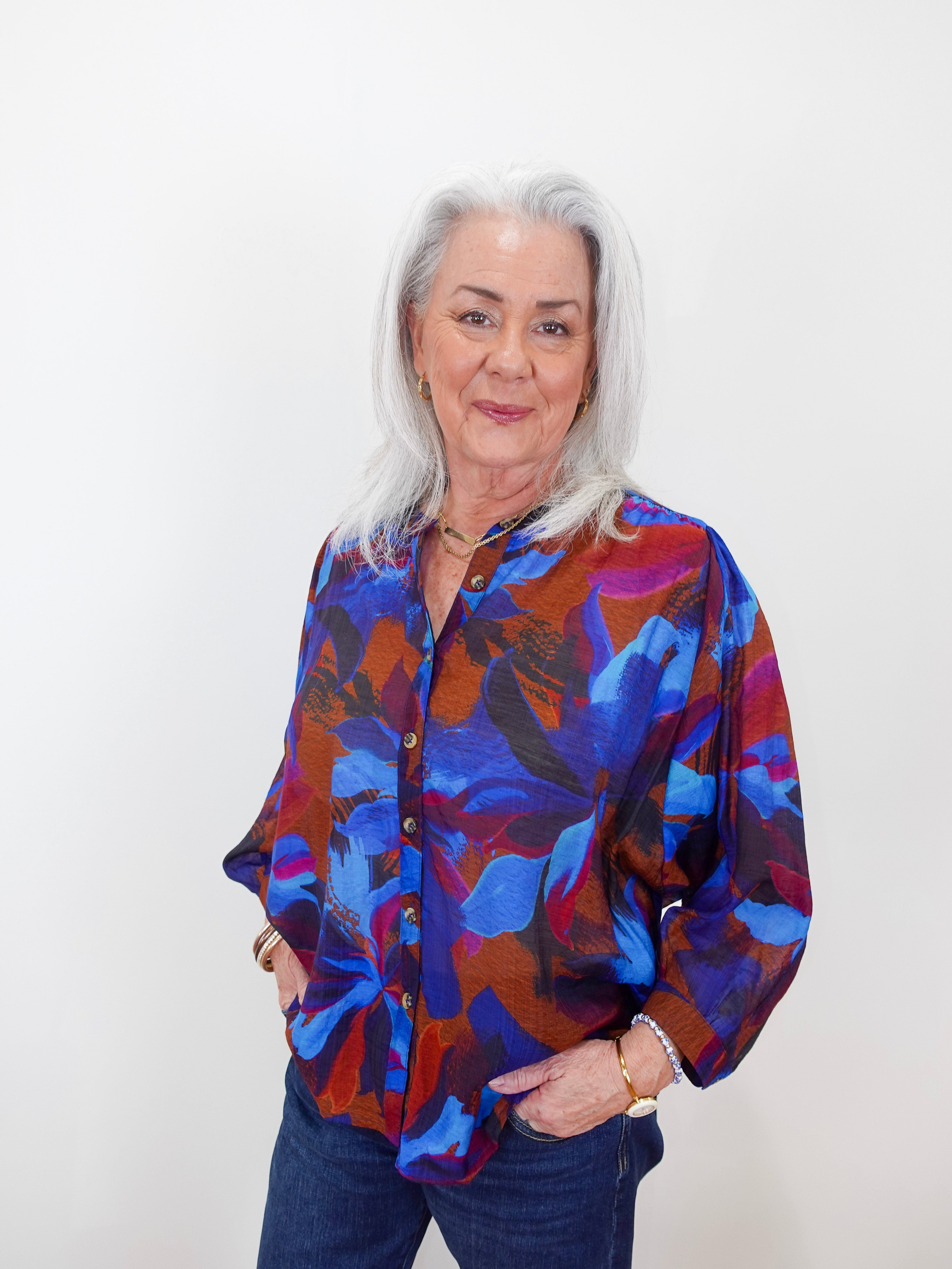 An older woman with silver hair stands against a plain white wall wearing a vibrant, long-sleeved button-up blouse with an abstract floral pattern in blue, purple, and red, paired with dark blue jeans.