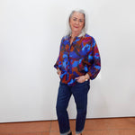 An older woman with white hair stands against a plain white wall wearing a colorful abstract print button-up shirt in blue, red, and purple, paired with dark blue jeans and black loafers.