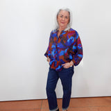 An older woman with white hair stands against a plain white wall wearing a colorful abstract print button-up shirt in blue, red, and purple, paired with dark blue jeans and black loafers.