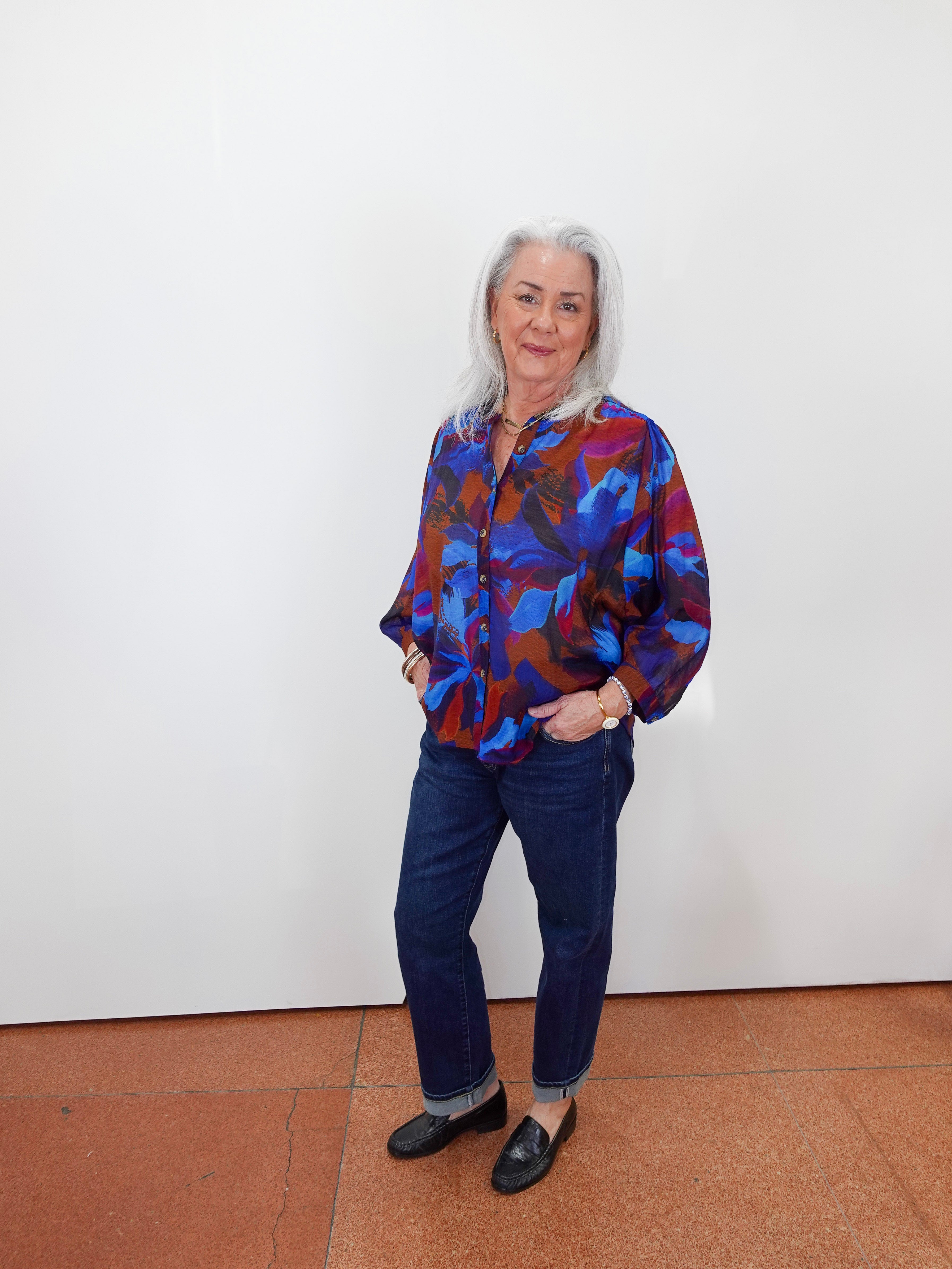 An older woman with white hair stands against a plain white wall wearing a colorful abstract print button-up shirt in blue, red, and purple, paired with dark blue jeans and black loafers.