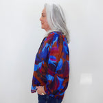 A woman with long gray hair is shown in profile wearing a vibrant abstract print blouse with blue, red, and purple tones, paired with dark blue jeans, against a plain white wall.