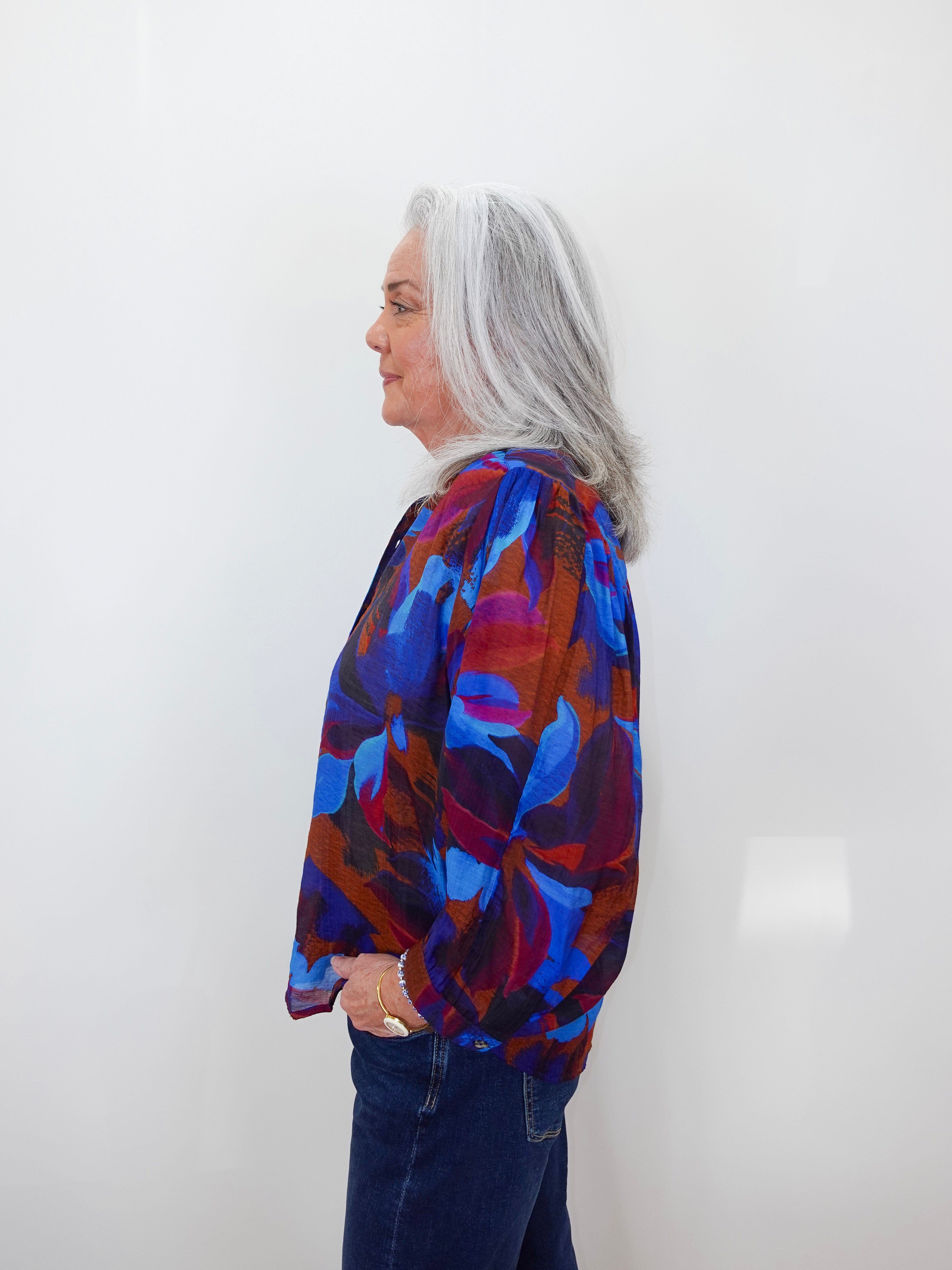 A woman with long gray hair is shown in profile wearing a vibrant abstract print blouse with blue, red, and purple tones, paired with dark blue jeans, against a plain white wall.