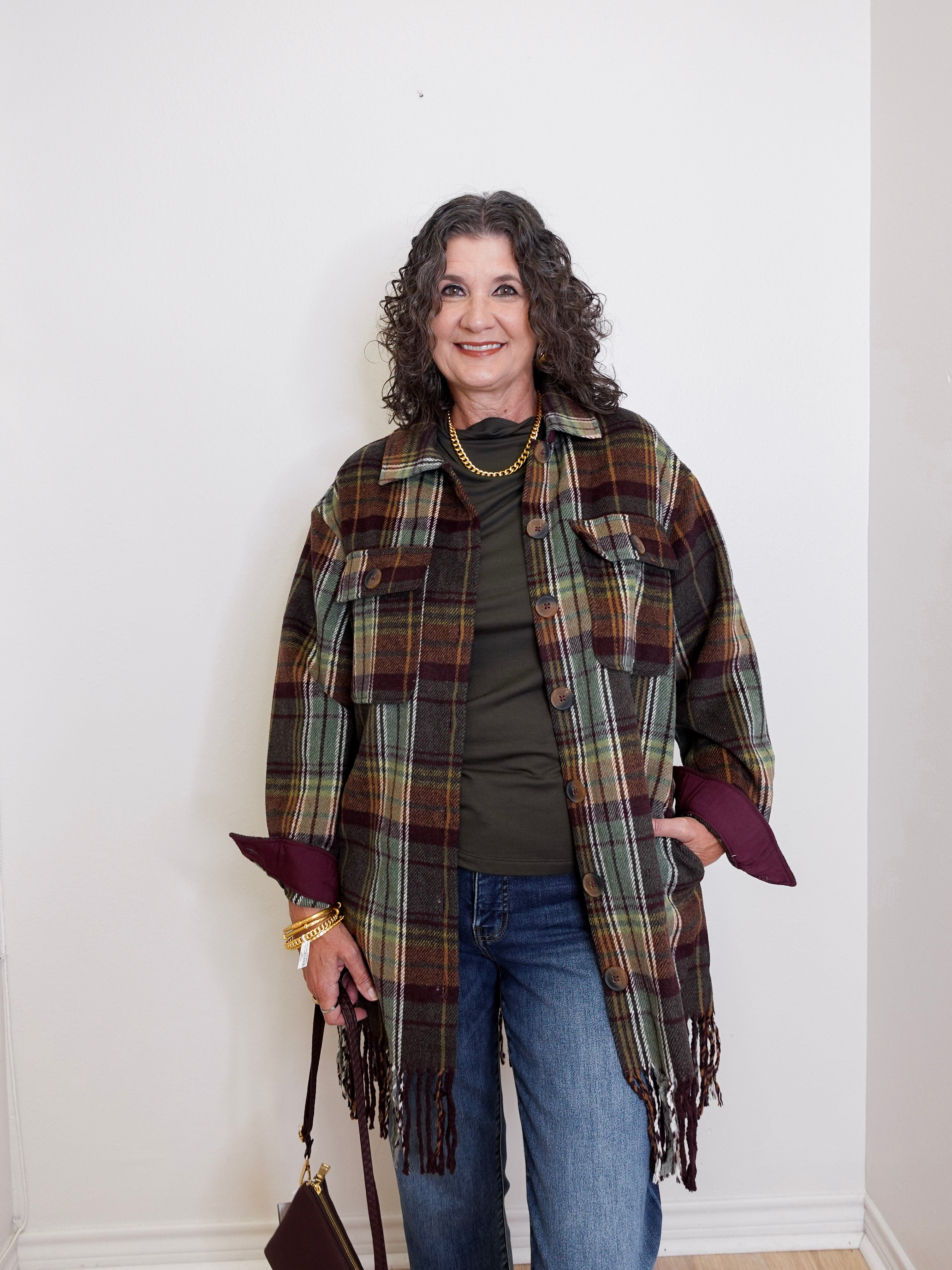Woman wearing a plaid shawl over a green top and blue jeans, standing against a white wall.