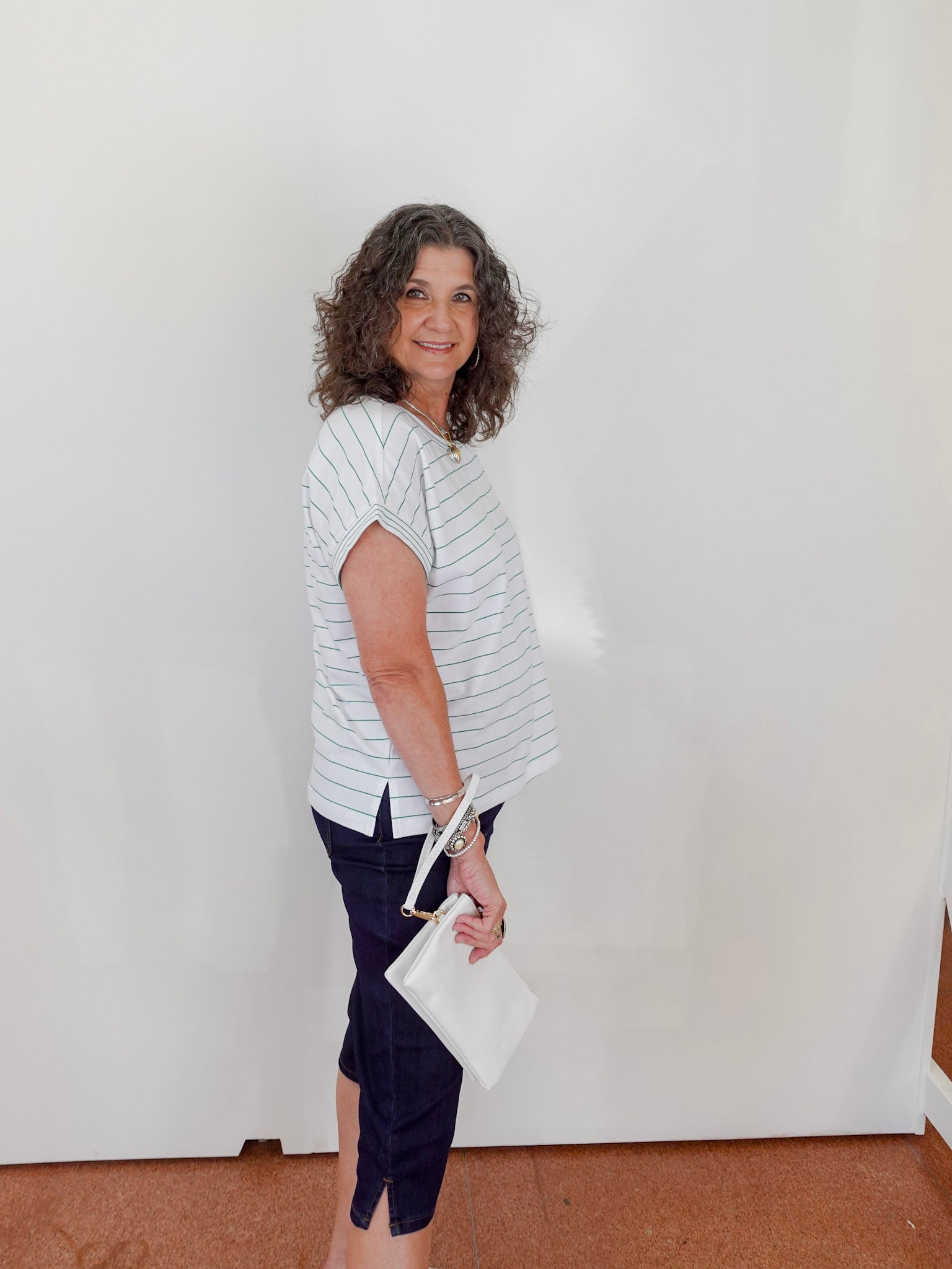 Woman wearing a striped shirt and dark pants holding a white clutch against a plain background