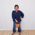 Woman in a blue top and jeans holding a brown handbag against a white wall.