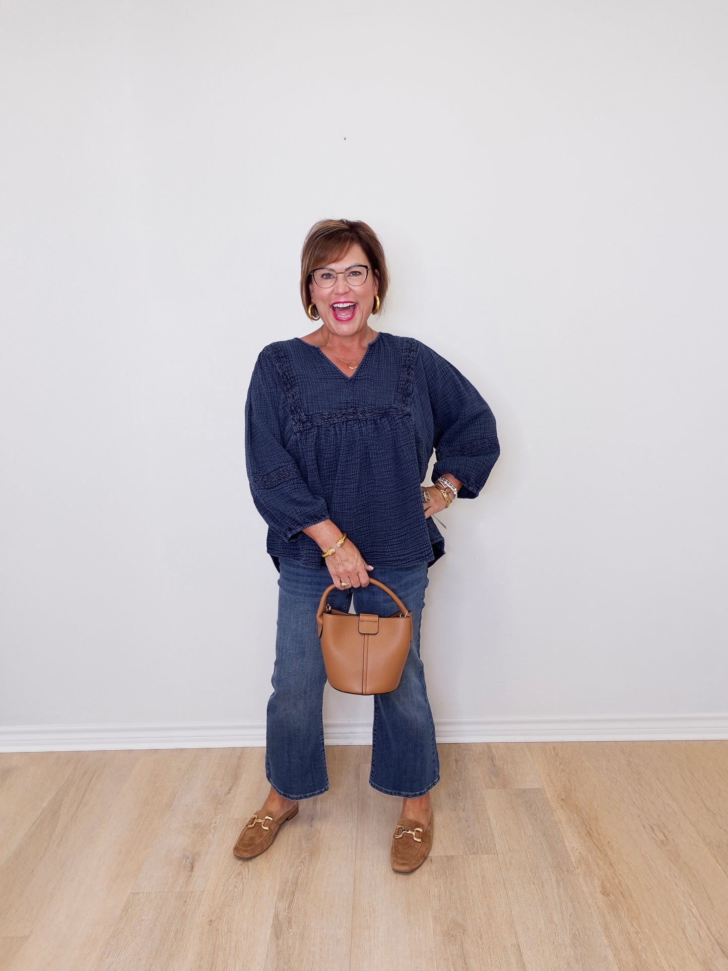 Woman in a blue top and jeans holding a brown handbag against a white wall.