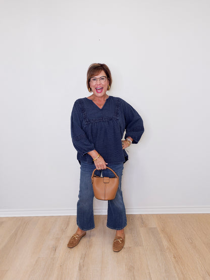 Woman in a blue top and jeans holding a brown handbag against a white wall.