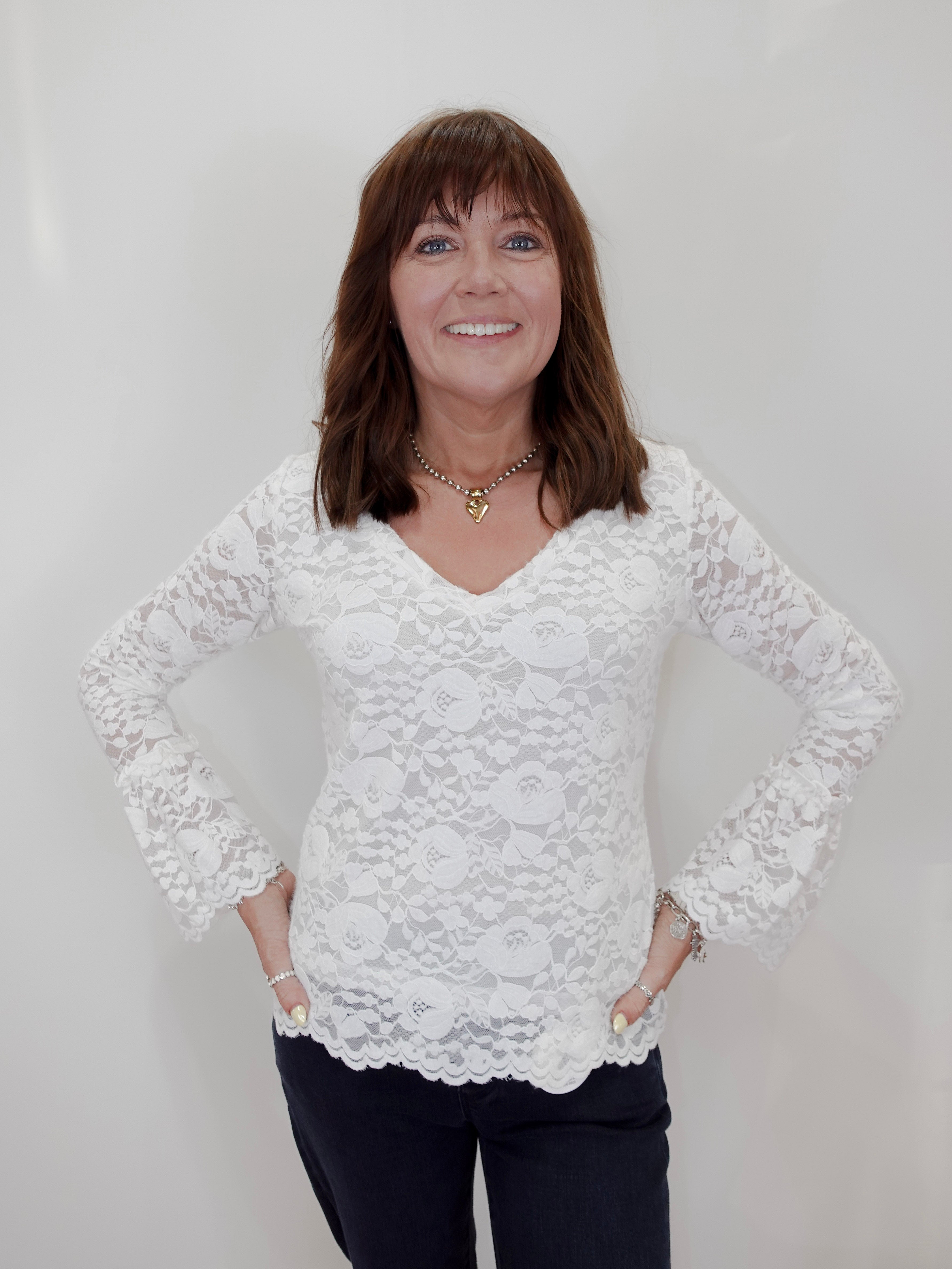 A woman with brown hair is smiling and posing with her hands on her hips, wearing a white lace long-sleeve blouse and dark pants, set against a plain white background.