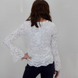 A woman shown from the back wearing a white lace long-sleeve top and dark jeans against a plain light gray wall.