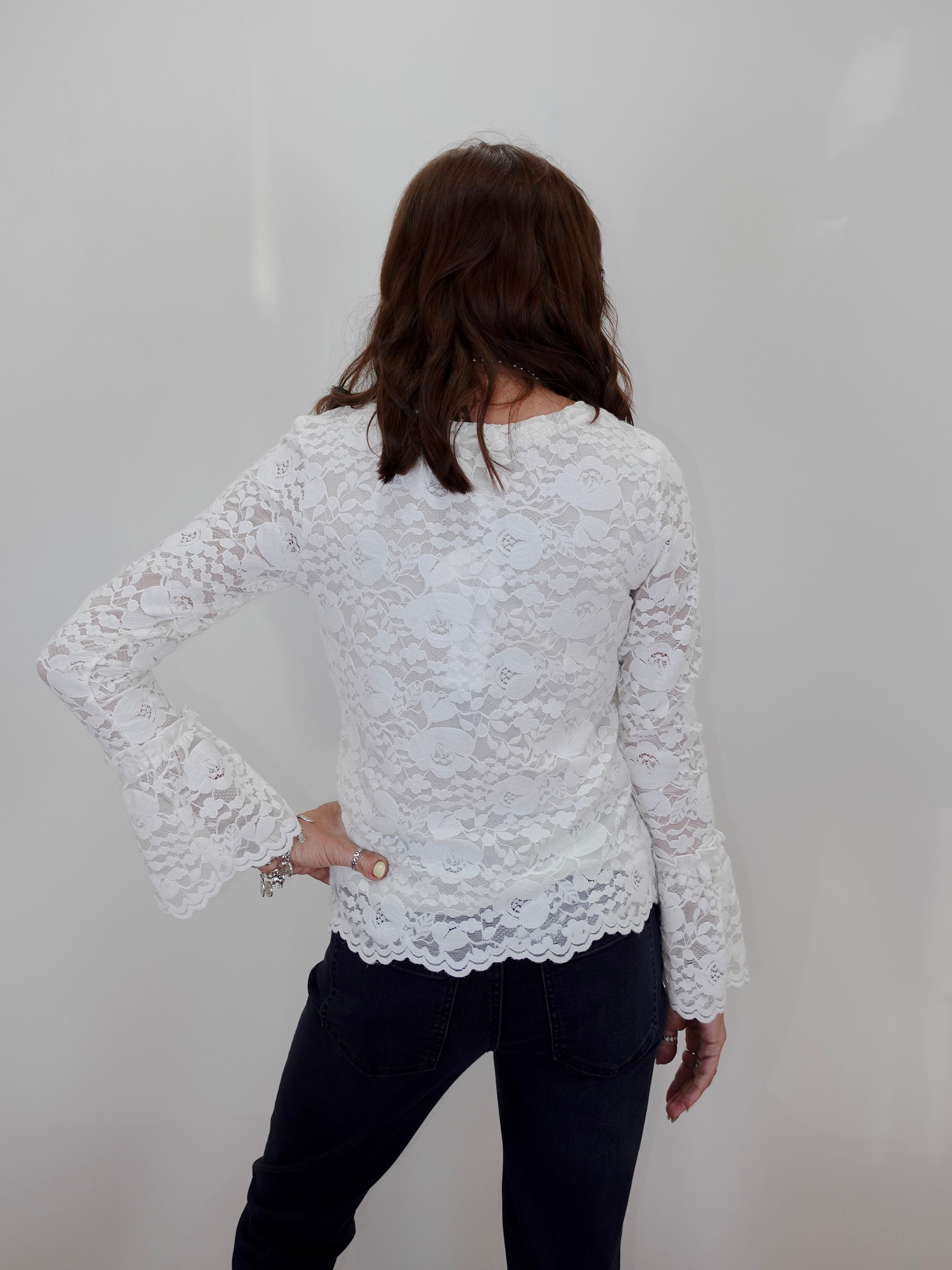 A woman shown from the back wearing a white lace long-sleeve top and dark jeans against a plain light gray wall.