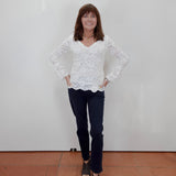 A woman stands against a plain white wall wearing a white lace long-sleeve top, dark navy pants, and black sandals.