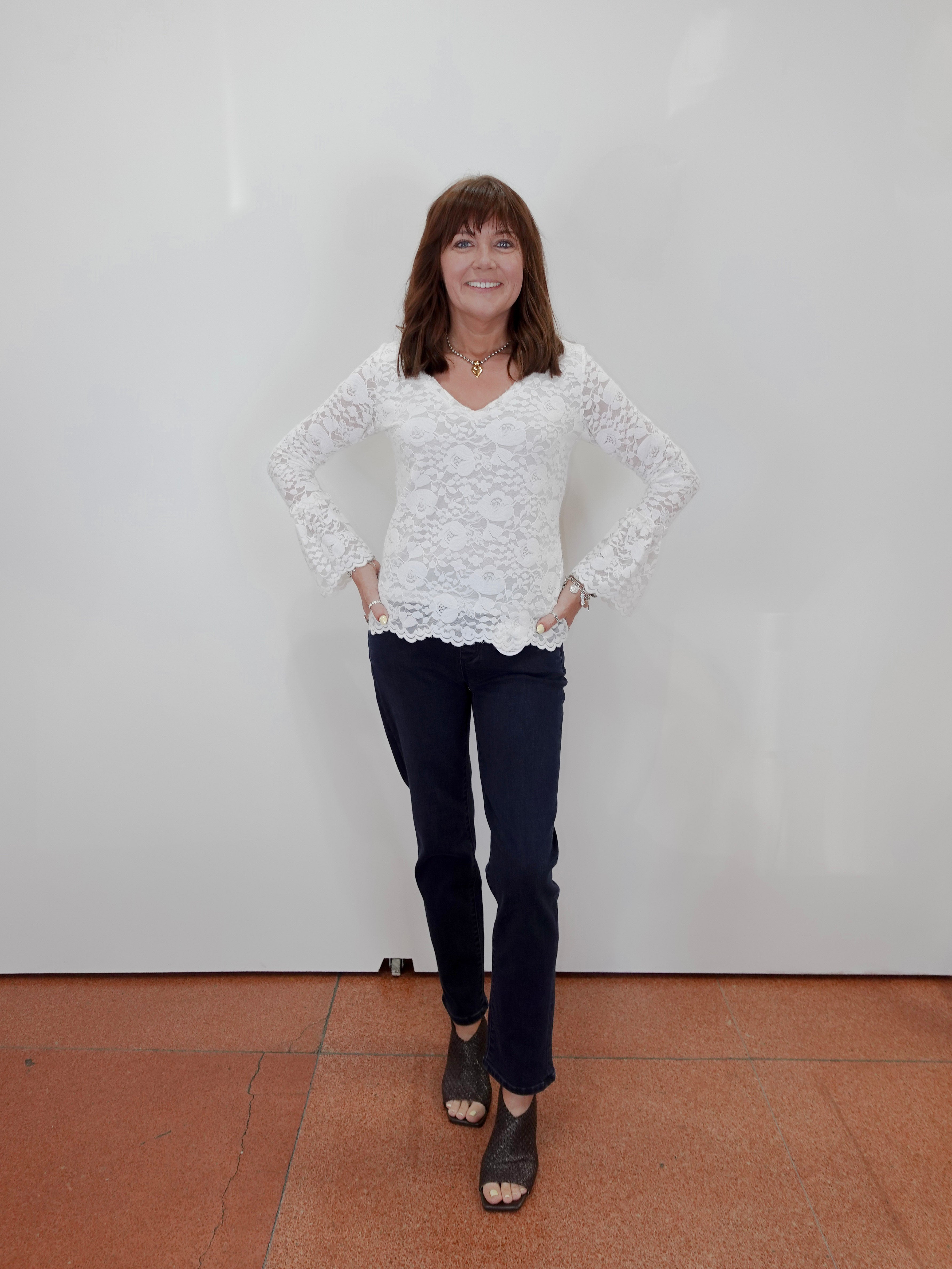 A woman stands against a plain white wall wearing a white lace long-sleeve top, dark navy pants, and black sandals.