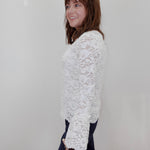 A woman with shoulder-length brown hair is shown from the side, wearing a white floral lace long-sleeve top and dark pants against a plain white wall.