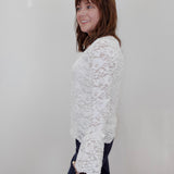 A woman with shoulder-length brown hair is shown from the side, wearing a white floral lace long-sleeve top and dark pants against a plain white wall.