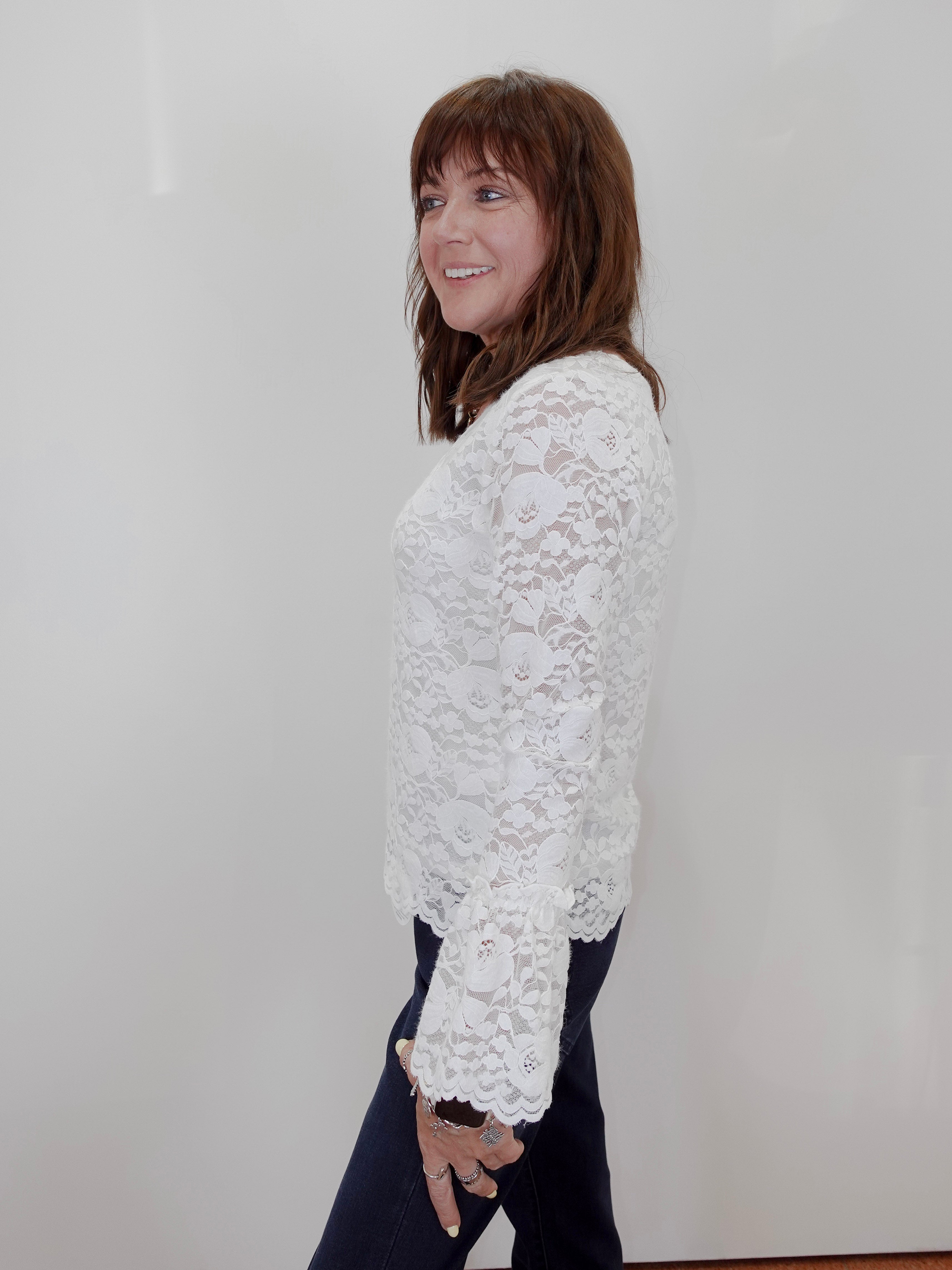 A woman with shoulder-length brown hair is shown from the side, wearing a white floral lace long-sleeve top and dark pants against a plain white wall.